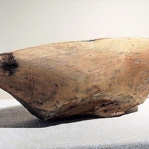 May include: A large, wooden, hand-carved bowl with a light brown color and a smooth, rounded surface. The bowl has a slightly pointed end and a wide, open mouth.