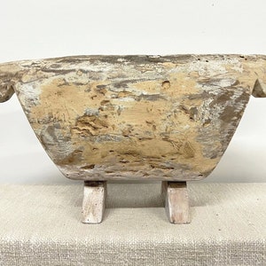 May include: A weathered, rectangular wooden bowl with a light brown and white finish. The bowl has rounded edges and is supported by two small, rectangular feet. The wood shows signs of age and wear, with visible grain and texture.