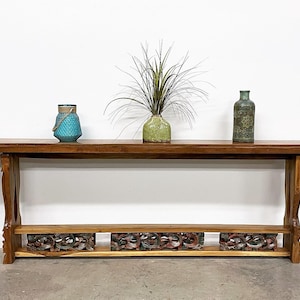 May include: A long, wooden console table with carved details on the legs and shelf. The table has a light brown finish and is decorated with three vases: a teal blue vase, a green vase with a plant, and a green glass bottle.