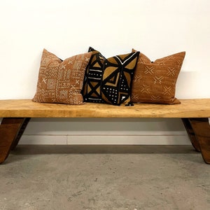 May include: A rustic wooden bench with a natural finish, featuring angled legs. Three decorative throw pillows in shades of brown and black rest on the bench. The pillows have patterned designs.