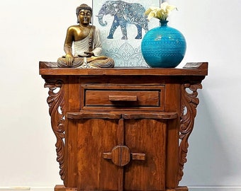 May include: A wooden cabinet with carved details, featuring a drawer and two doors. A golden Buddha statue sits on top of the cabinet, along with a blue vase and a framed print of an elephant.