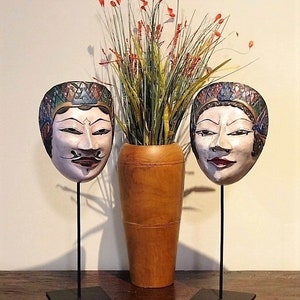 Puede incluir: Dos máscaras de madera coloridas con diseños intrincados en soportes negros, un jarrón con follaje verde y rojo en el centro.