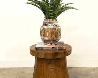 May include: A wooden side table with a round top and a wide, cylindrical base. A book titled "Dame Traveler" is on top of the table, and a white ceramic vase with a fern plant is on top of the book.