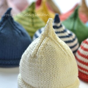 May include: A collection of knitted baby hats in various colors, including white, blue, green, and red. The hats are all made with a simple stitch pattern and have a pointed top. The text "rocket clothing london" is visible in the image.
