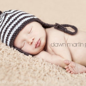 Pode incluir: Um recém-nascido usando um gorro de tricô listrado marrom e branco com uma cauda longa, dormindo em um cobertor bege felpudo.