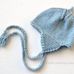 May include: A light blue knitted baby hat with a long, braided tie. The hat has a pointed top and a small tassel on the end of the tie.