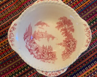 May include: A white oval serving dish with a pink floral design. The design features a pink castle, trees, and flowers. The dish has a scalloped edge and two handles.