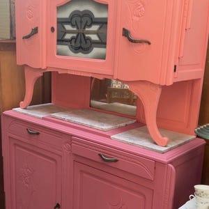 Könnte beinhalten: Ein zweiteiliger antiker Schrank, der in einem leuchtenden Korallenrosa lackiert ist. Der obere Teil hat eine Glastür mit einem dekorativen Design, während der untere Teil zwei Türen und marmorierte Oberflächen hat. Der Schrank hat verzierte Schnitzereien und dunkle Griffe.