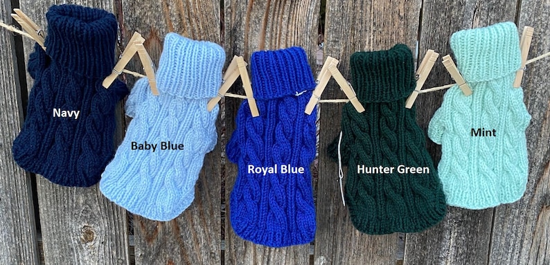 May include: Five knitted dog sweaters hanging on a clothesline. The sweaters are in the colours navy, baby blue, royal blue, hunter green, and mint.