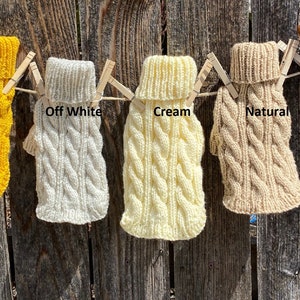 May include: Five knitted dog sweaters in different colours hanging on a clothesline. The sweaters are labelled "Gold", "Off White", "Cream", "Natural", and "Copper".