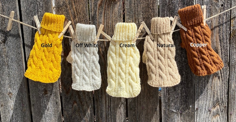 May include: Five knitted dog sweaters in different colours hanging on a clothesline. The sweaters are labelled "Gold", "Off White", "Cream", "Natural", and "Copper".