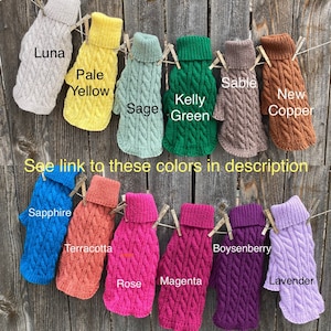 May include: A display of 12 different coloured knitted dog sweaters hanging on a wooden fence. The sweaters are all the same style, with a cable knit pattern and a turtleneck. The colours are: Luna, Pale Yellow, Sage, Kelly Green, Sable, New Copper, Sapphire, Terracotta, Rose, Magenta, Boysenberry, and Lavender.