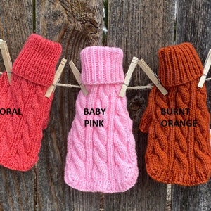 May include: Five knitted dog sweaters in different colours hanging on a clothesline. The sweaters are red, coral, baby pink, burnt orange, and hot pink. Each sweater has a turtleneck and a cable knit pattern.