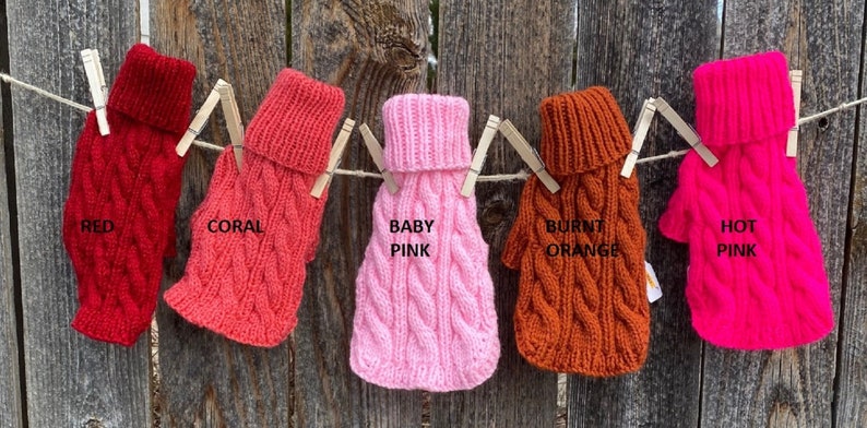 May include: Five knitted dog sweaters in different colours hanging on a clothesline. The sweaters are red, coral, baby pink, burnt orange, and hot pink. Each sweater has a turtleneck and a cable knit pattern.