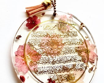 May include: A round, clear resin ornament with gold lettering and pressed pink and white flowers. The ornament is hanging from a gold chain with a pink tassel.