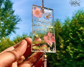 May include: A rectangular resin charm with a gold tassel. The charm features pressed flowers and gold leaf, and includes an Arabic inscription in gold lettering. The inscription reads: "Subhana alladhi sakhkhara lana hadha wa ma kunna lahu muqrinin. Wa inna ila rabbina la munqalibun."
