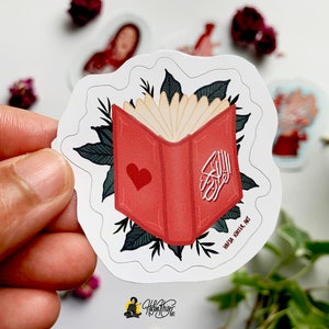 May include: A red book with a heart on the cover and Arabic calligraphy. The book is surrounded by green leaves. The text "Haya Khila Art" is at the bottom of the sticker.