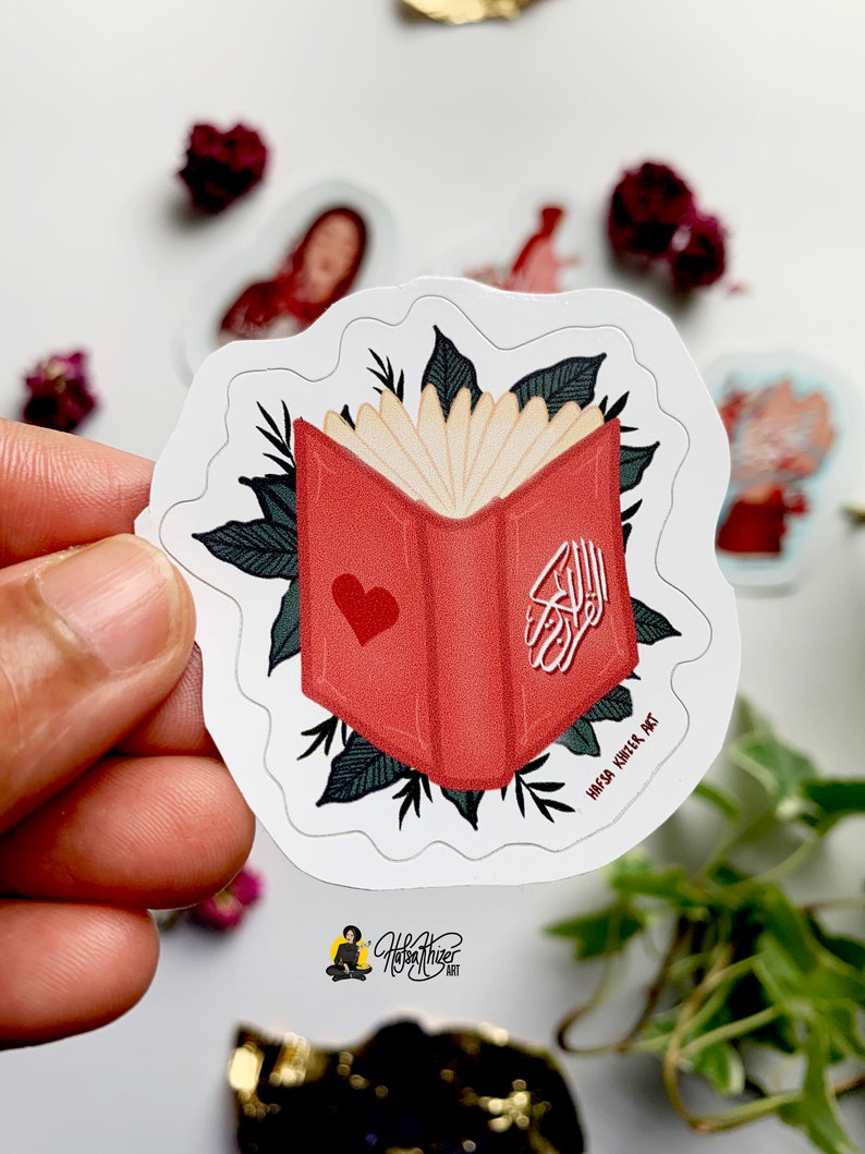 May include: A red book with a heart on the cover and Arabic calligraphy. The book is surrounded by green leaves. The text "Haya Khila Art" is at the bottom of the sticker.