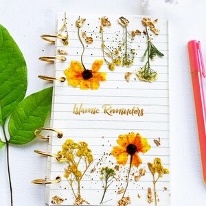 May include: A clear, lined notebook with gold spiral binding, featuring pressed yellow flowers and gold leaf accents. The words "Islamic Reminders" are written in gold script. A pink pen rests on the side.