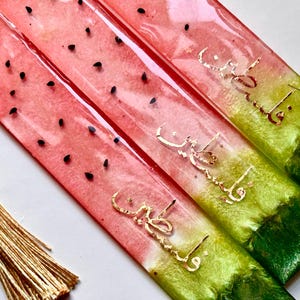 May include: Three watermelon-themed bookmarks with gold calligraphy and a tassel. The bookmarks are made of clear resin with a red and green gradient. The calligraphy is in Arabic.