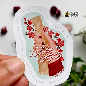 May include: A sticker with a hand holding a rope with the Arabic text "Wa Inna laka la ma'a al-husni", which translates to "And indeed, for you is with the good", in red and black calligraphy. The background is a light blue with pink cherry blossoms.