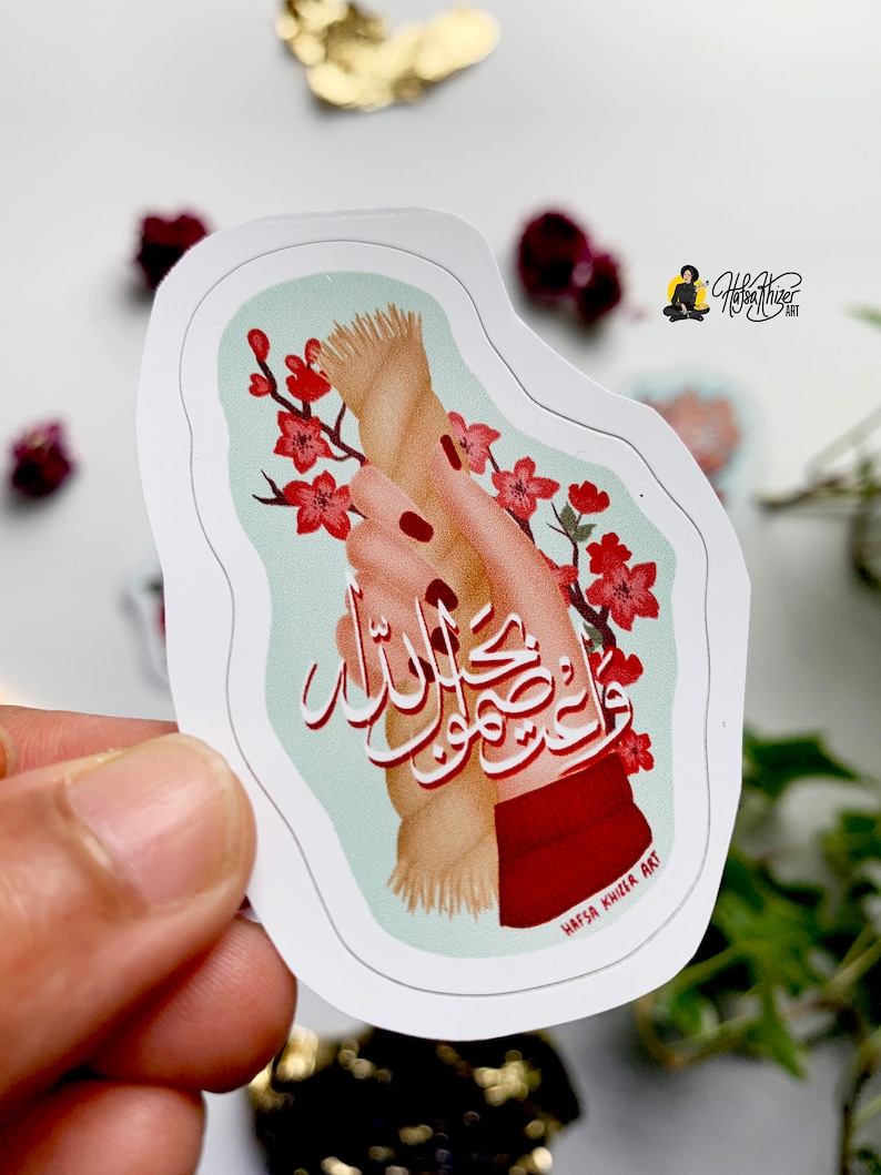 May include: A sticker with a hand holding a rope with the Arabic text "Wa Inna laka la ma'a al-husni", which translates to "And indeed, for you is with the good", in red and black calligraphy. The background is a light blue with pink cherry blossoms.
