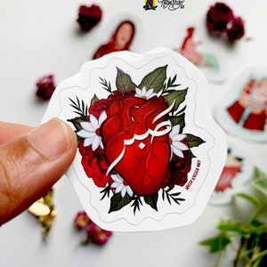 May include: A red anatomical heart with Arabic calligraphy inside, surrounded by white and red flowers and green leaves. The sticker has a white border and the text "Haifa Khnezer Art" at the bottom.