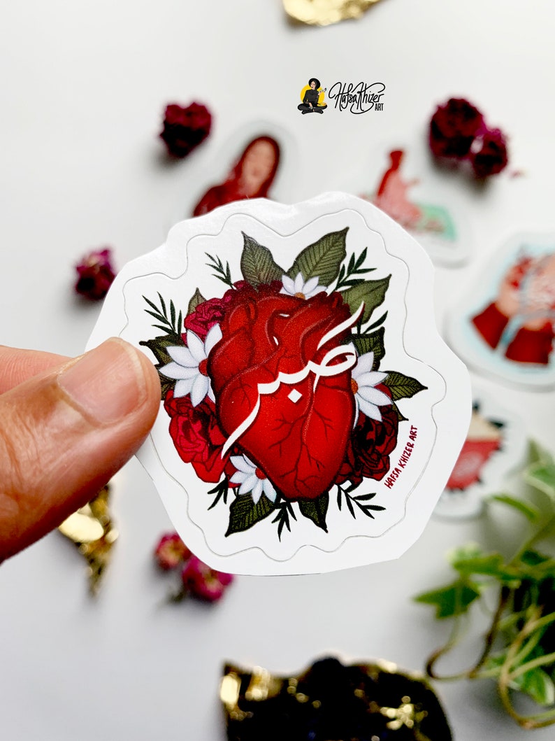 May include: A red anatomical heart with Arabic calligraphy inside, surrounded by white and red flowers and green leaves. The sticker has a white border and the text "Haifa Khnezer Art" at the bottom.