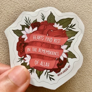 May include: A red heart surrounded by red and white flowers with green leaves. The heart has a red ribbon wrapped around it with the text "Hearts find rest in the remembrance of Allah". The sticker is designed by Hafsa Khizer Art.