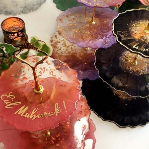 May include: A set of three tiered cake stands with gold accents. The stands are decorated with different colours and patterns, including purple, green, black, and red. The top tier has the words "Eid Mubarak!" written in gold calligraphy.