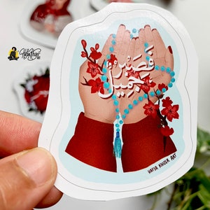 May include: A sticker with a blue and white prayer beads in two hands. The hands are surrounded by red flowers and the Arabic text "Wa Inna La Rabbi Na Shkur" is written in white. The sticker has the artist's name "Hafsa Khilzi Art" at the bottom.