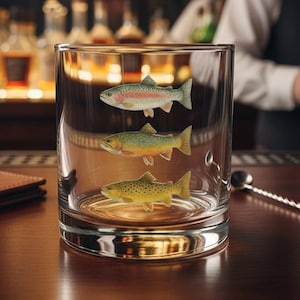 May include: A clear glass tumbler featuring three distinct fish illustrations stacked vertically on the exterior surface. The glass has a heavy base and smooth, straight sides, showcasing detailed, colorful depictions of a rainbow trout, a brook trout, and a brown trout.