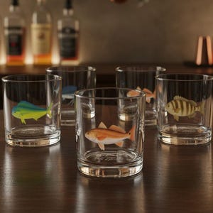 May include: Six clear, round drinking glasses with different colorful fish illustrations. The fish include a yellow and blue mahi-mahi, a blue and white fish, an orange fish, a striped fish, and two other fish. The glasses are on a dark wooden surface.