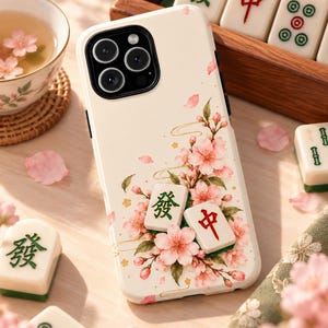 Puede incluir: Una funda de smartphone color crema con un diseño estampado de flores de cerezo rosas y dos fichas de mahjong tridimensionales con caracteres verdes y rojos. La funda tiene un recorte negro para la lente de la cámara.