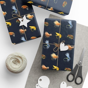May include: Gift-wrapped presents with a navy blue background and a repeating pattern of colorful fishing flies. The gifts are tied with twine and adorned with white star and heart-shaped tags. A spool of twine, scissors, and blank tags are also visible.