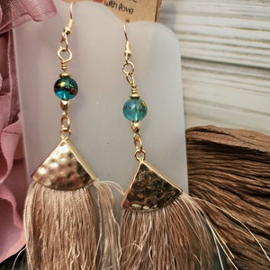 Puede incluir: Pendientes de tono dorado con un colgante triangular de metal martillado y una cuenta de vidrio azul turquesa. Los pendientes tienen un borla beige.