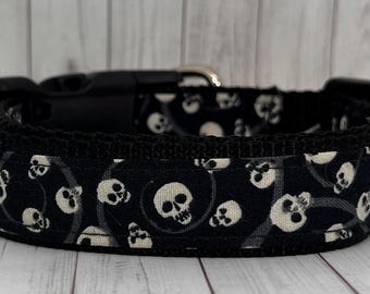 May include: Black dog collar featuring a repeating pattern of white skulls on a black background. The collar has a black plastic buckle and a metal loop for leash attachment. The collar is made of durable fabric.