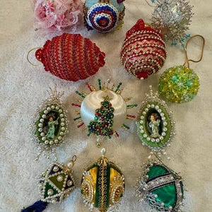 May include: A collection of twelve decorative Christmas ornaments featuring various designs, including beaded spheres, fabric-covered shapes, and oval ornaments with small angel figurines inside, adorned with sequins, lace, and metallic trim.
