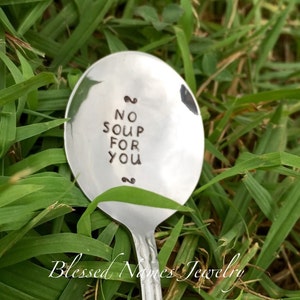 May include: A silver spoon with the text "NO SOUP FOR YOU" stamped on the bowl. The spoon is set against a backdrop of green grass. The words "Blessed Names Jewelry" are at the bottom of the image.