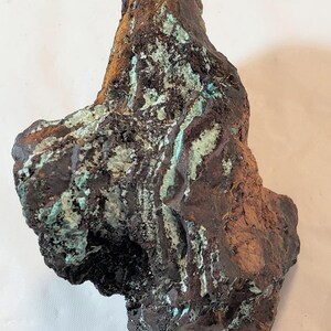 Puede incluir: Una roca rugosa de forma irregular con una gama de colores marrón oscuro, negro y verde. La roca tiene una superficie rugosa y texturizada y parece ser una muestra mineral natural.