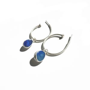 May include: A pair of silver hoop earrings with a blue gemstone pendant. The gemstones have a rounded shape and are set in a silver bezel. The earrings are displayed on a white background.