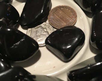 May include: A white ceramic dish filled with black obsidian stones. Two US coins, a penny and a nickel, are placed among the stones.
