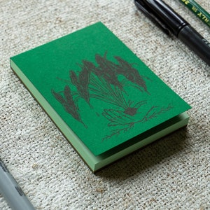 May include: A small, green notepad with a dark illustration of a plant and hand design. The notepad is open, revealing the pages. A black permanent marker, a green pencil, and a gray marker are in the background.