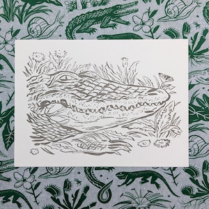 May include: A black and white line art print of an alligator's head emerging from a swamp. The print is on a white background.