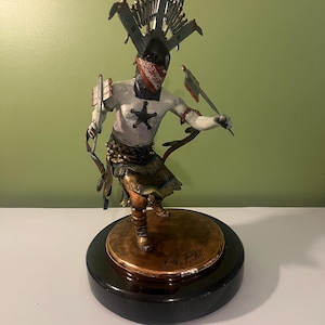 May include: Bronze sculpture of a Native American figure in a dynamic pose, wearing a headdress, bandana, and star badge. The figure holds weapons and is mounted on a round base with a black marble pedestal. The sculpture is signed.