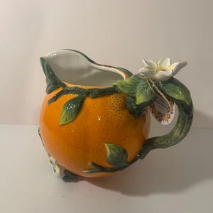 May include: A ceramic pitcher shaped like a textured orange with a glossy finish. The handle is designed as a green branch with sculpted green leaves and a white orange blossom flower. A small paper tag is attached to the handle with twine.