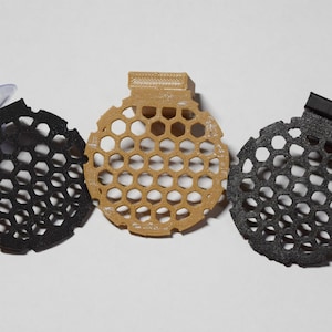 May include: Three round, black and beige, honeycomb-shaped air fresheners. Two of the air fresheners have a suction cup and a hook on the back. The third air freshener has a hook on the back.