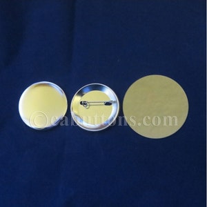 May include: Three blank, round pinback buttons. One button is face up, showing a shiny, gold-colored surface. Another button shows the back with a safety pin. A third button is a gold-colored, round paper backing. The background is a dark blue fabric.