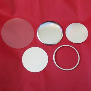 May include: Five round, blank button blanks for making custom buttons. The blanks are in different sizes and materials, including clear plastic, white plastic, silver metal, and a silver metal ring.