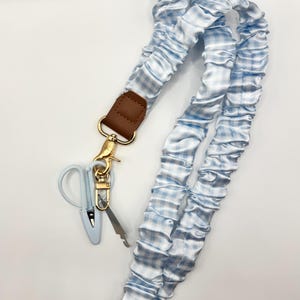 May include: A light blue and white gingham patterned fabric lanyard with a gathered, scrunchie-style texture. It features a brown faux leather connector, a gold-toned metal lobster clasp, and a small light blue plastic scissor-shaped charm.